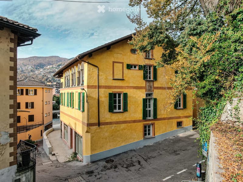 A yellow multi-story building with green shutters and a balcony, situated on a narrow street with a view of a hilly landscape in the background.