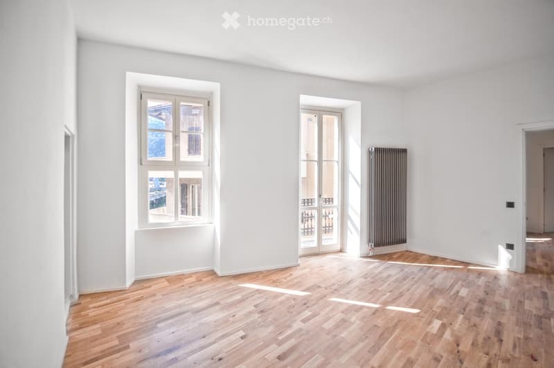 Empty room with light wooden flooring, white walls, large windows, and a radiator. No furniture or equipment present.