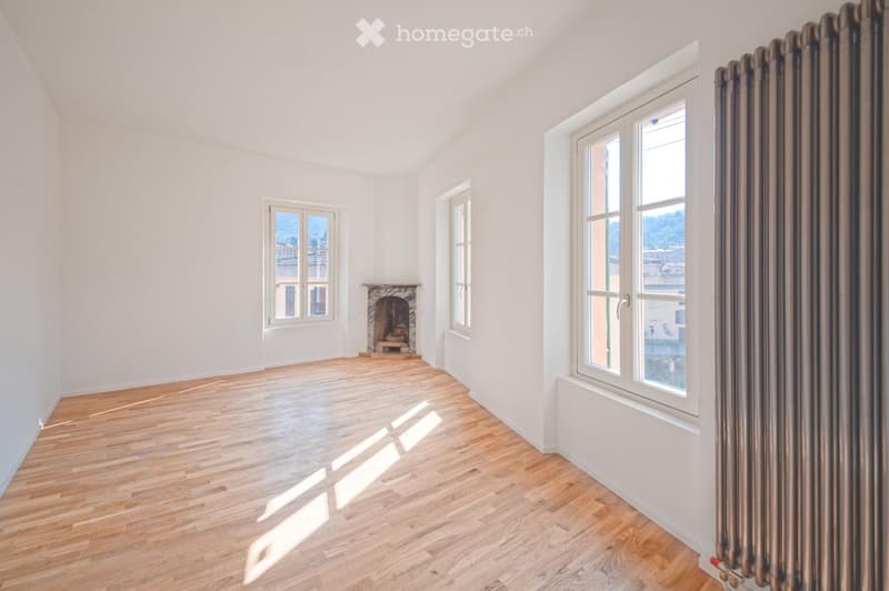 Empty room with wooden flooring, white walls, and large windows. Features a fireplace and natural light streaming in.