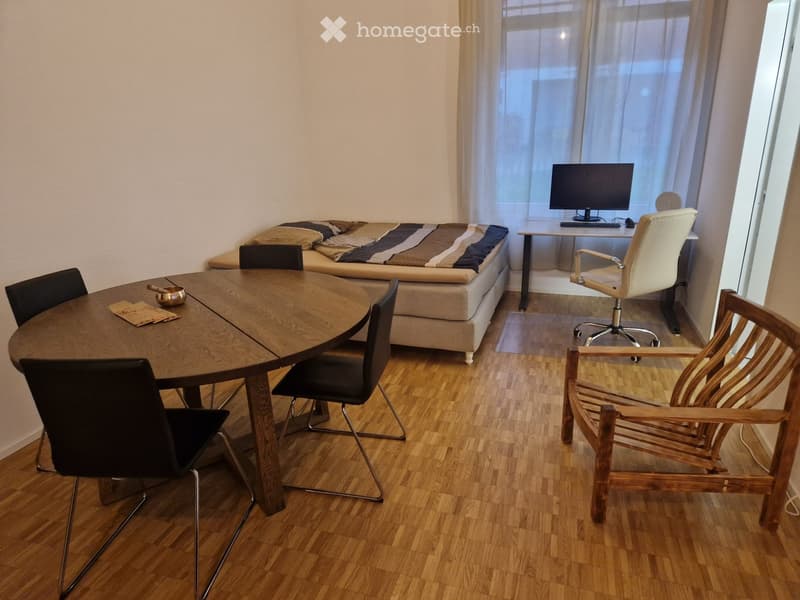 A furnished room with a round wooden table, chairs, a sofa, a laptop, and a screen. The room has wooden flooring and large windows with curtains.