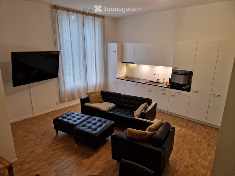 Modern living room with a black leather sofa, ottoman, armchair, and a flat-screen TV. The room features a kitchen area with white cabinets, a sink, and an oven. The space is well-lit with large windows and wooden flooring.