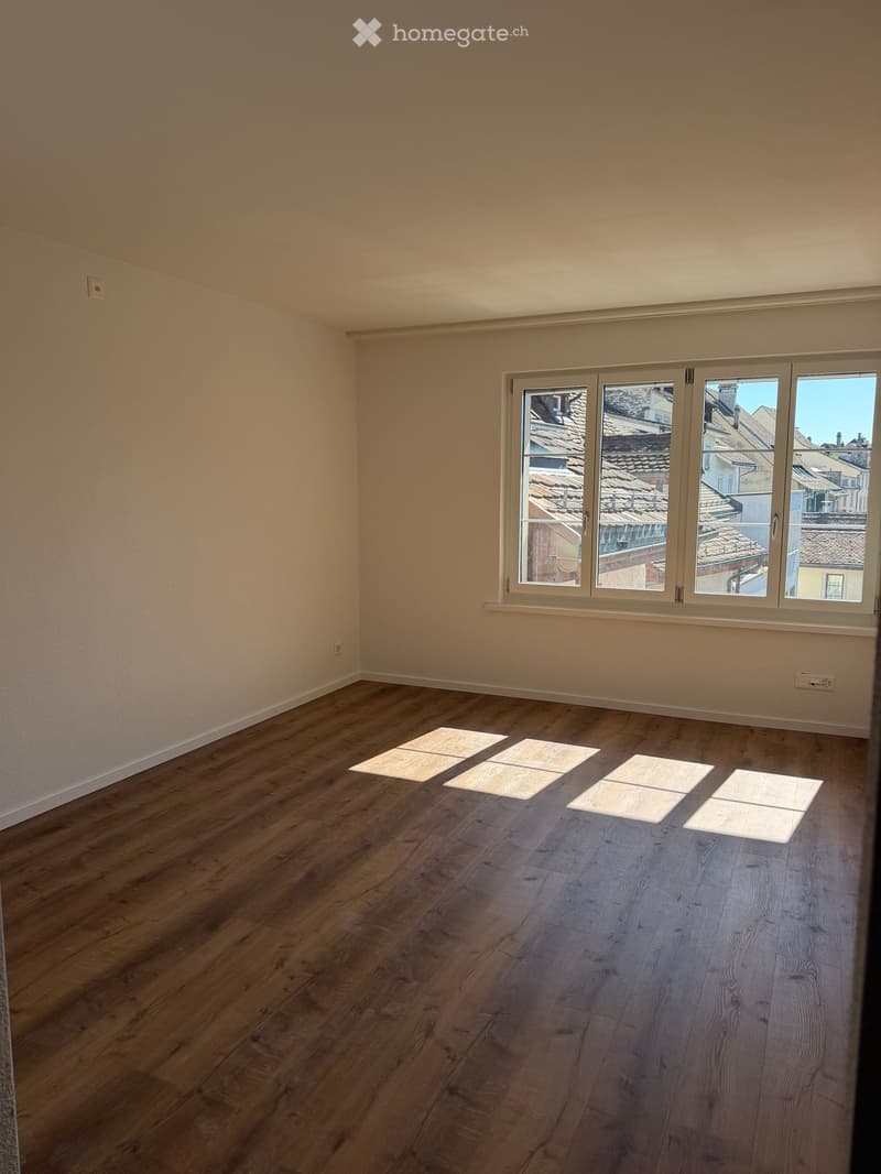 Empty room with wooden flooring and large windows showing a view of rooftops. No furniture or equipment present.