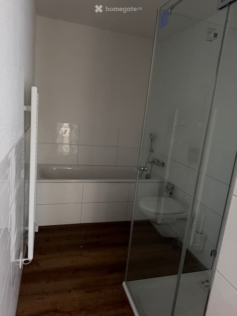 Modern bathroom with a bathtub, toilet, and glass-enclosed shower. The space features light grey tiles and a wooden floor.
