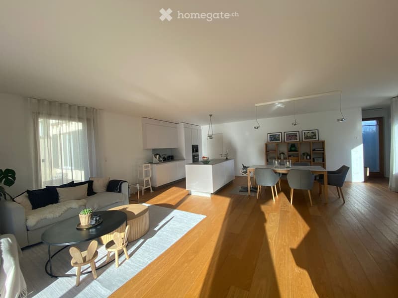 Modern open-plan living and dining area with light wood flooring, white kitchen cabinets, a grey sofa with cushions, a round coffee table, an ottoman, and a dining table with chairs.