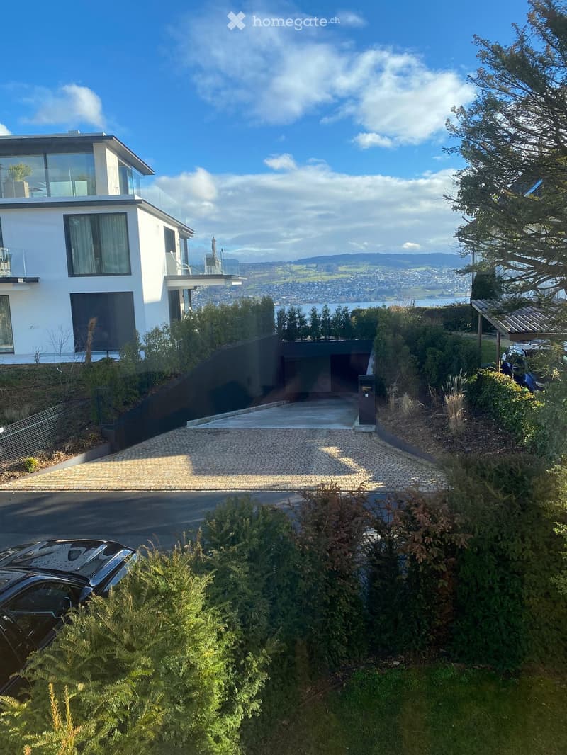 Modern two-story house with white exterior, large windows, and a balcony, situated on a hillside with a scenic view of the city and water.