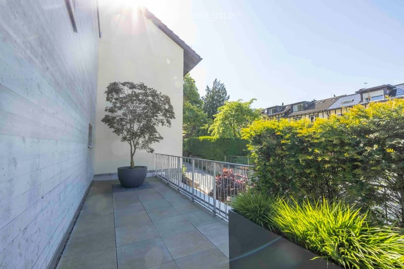 A modern balcony with tiled flooring, a metal railing, and potted plants, including a small tree and shrubs, overlooking a green area with trees and neighboring buildings.