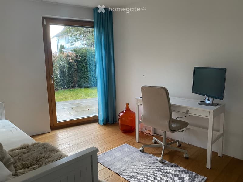 Cozy bedroom with a bed, desk, chair, and a large window with blue curtains. The room features a wooden floor, a striped rug, and a decorative vase.