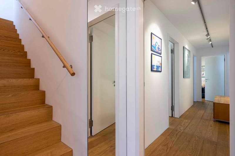 Modern hallway with wooden staircase, white walls, and recessed lighting. Features framed artwork and open doors leading to other rooms.