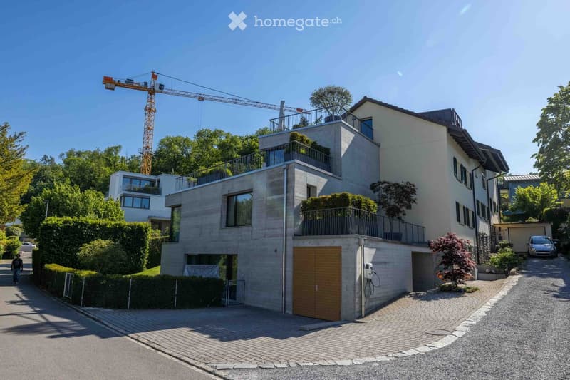 Modern two-story house with a contemporary design, featuring large windows, balconies, and a wooden garage door. The house is surrounded by greenery and has a paved driveway.