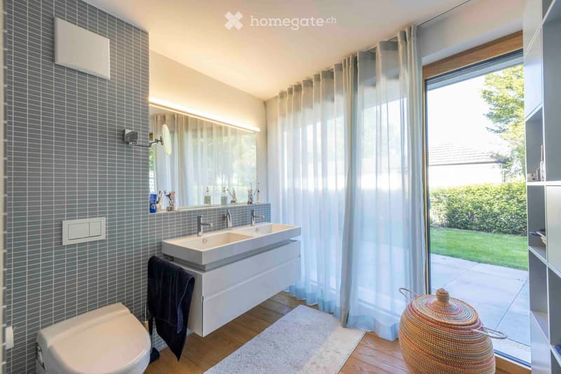 Modern bathroom with double sink, toilet, large mirror, and wicker basket near the window. The room features grey tiled walls, wooden flooring, and sheer curtains.
