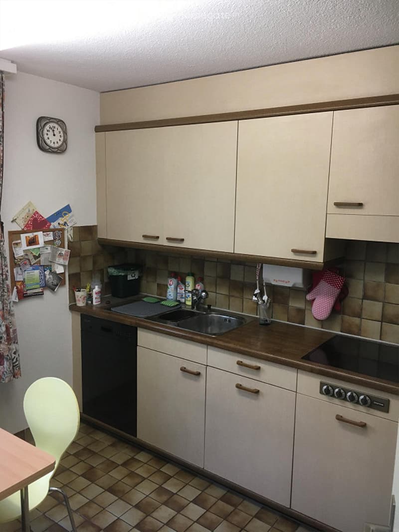 A modern kitchen with light-colored cabinets, a wooden countertop, a sink, a faucet, an oven, a dishwasher, and a chair. The walls are tiled, and there is a clock on the wall.