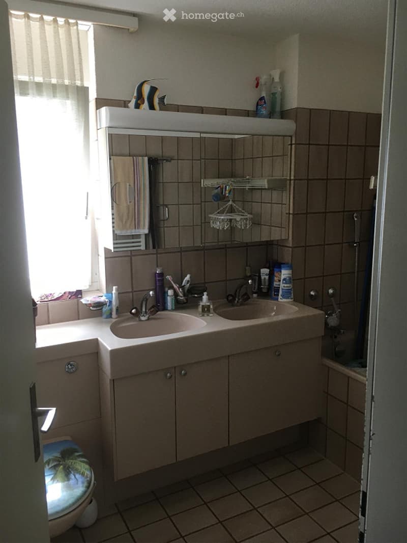 A small bathroom with a double sink vanity, mirror, tiled walls and floor, and a toilet. The vanity has a towel hanging on the side and various toiletries on the counter.