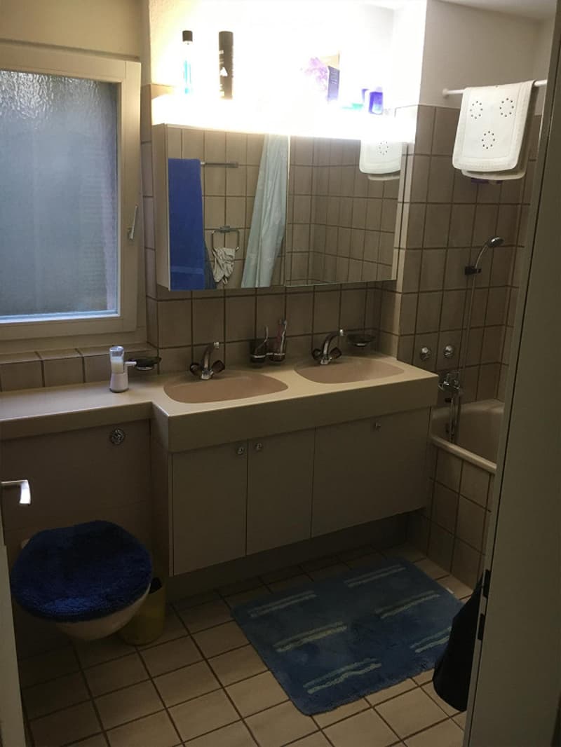 A modern bathroom with beige tiles, a double sink vanity, a large mirror, a toilet, a bathtub, and a shower area. The space includes a blue towel, a toothbrush holder, and a shower head.