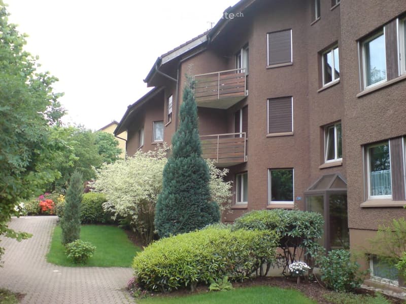 Multi-story apartment building with brown facade, multiple windows, and a balcony on the upper floor. The surrounding area includes a well-maintained garden with various shrubs, trees, and a paved pathway.