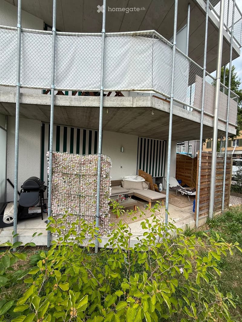 Outdoor patio area with a sofa, coffee table, lounge chairs, and a grill, partially enclosed by a metal fence and surrounded by greenery.