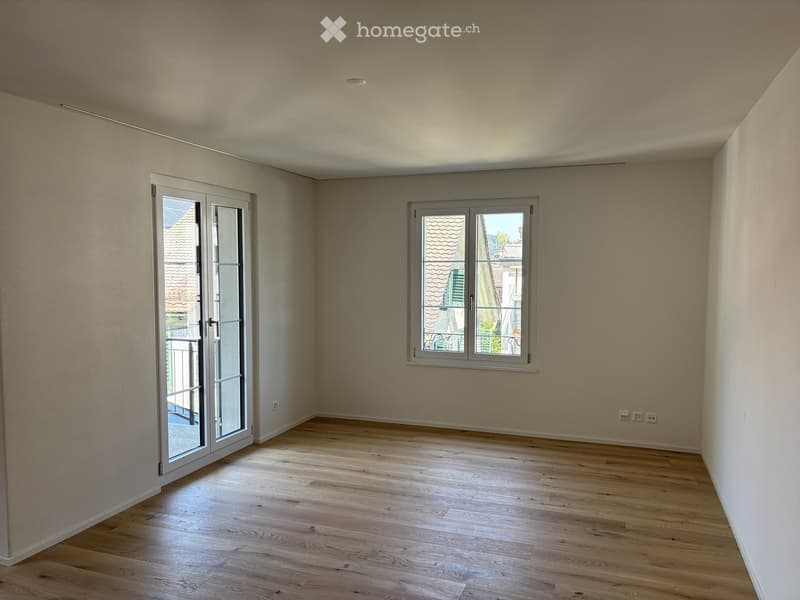 Empty room with light wooden flooring, white walls, and large windows, including a sliding glass door leading to a balcony.