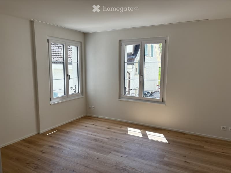 Empty room with light wooden flooring and white walls, featuring two large windows with white frames and metal railings visible outside.