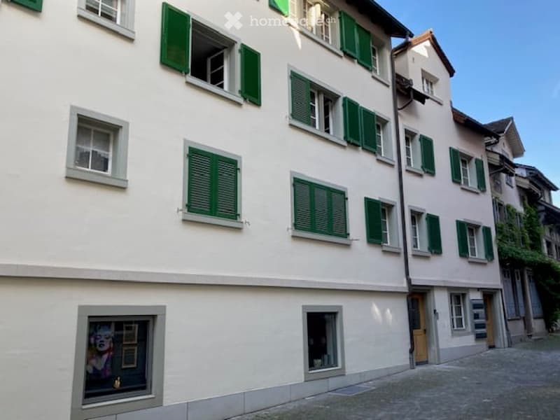 A multi-story residential building with white walls, green shutters, and multiple windows. The building features a mix of architectural styles and is situated in a cobblestone street setting.