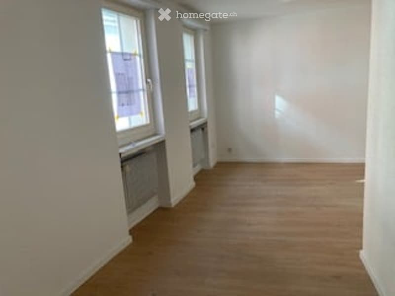 Completely empty room with wooden flooring, white walls, and large windows allowing natural light to enter.