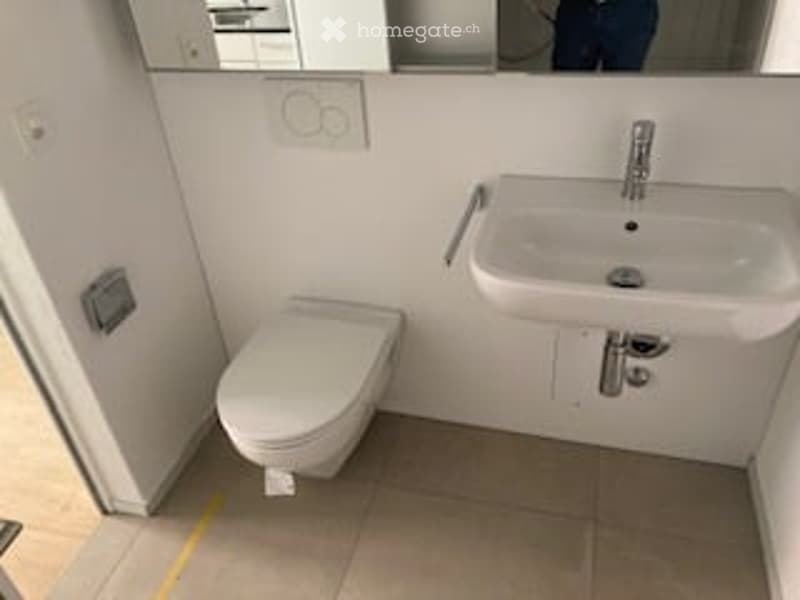 Modern, compact bathroom with a white toilet and a square sink with a chrome faucet. The walls are white, and the floor is tiled in a light brown color.