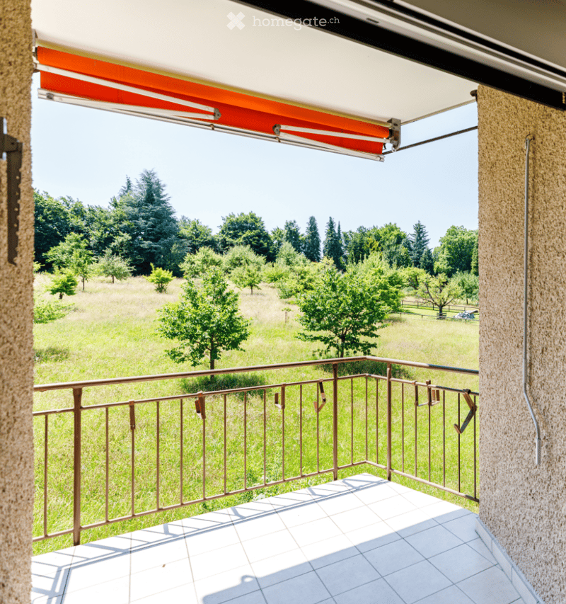 Balcony with tiled floor, metal railing, and retractable awning, overlooking a green field with trees.