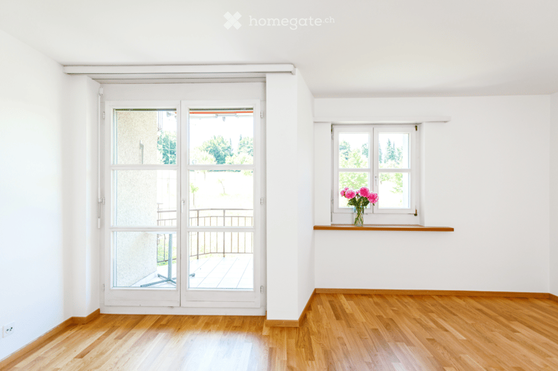 Empty room with wooden flooring, white walls, large windows, and a balcony door leading to a balcony.
