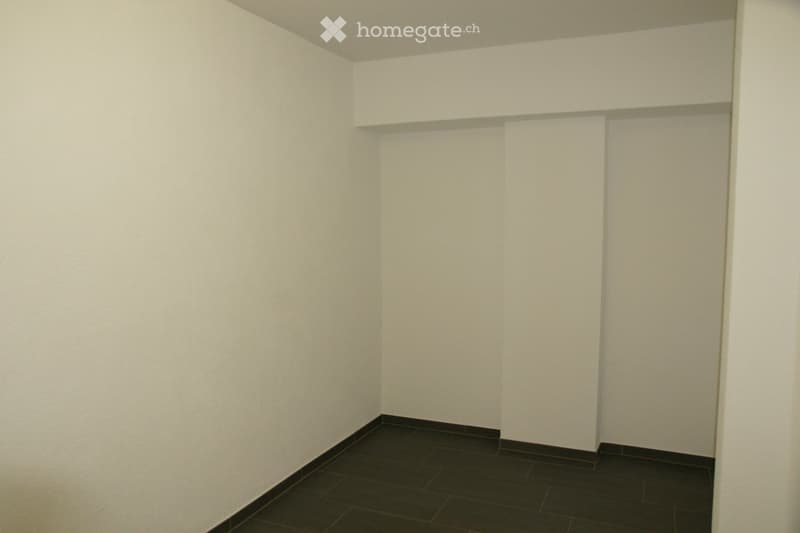 Completely empty room with bare white walls and dark tiled floor. No furniture or equipment.