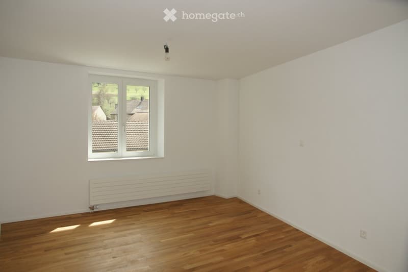 Empty room with wooden flooring, white walls, a large window with white frame, and a radiator below the window.