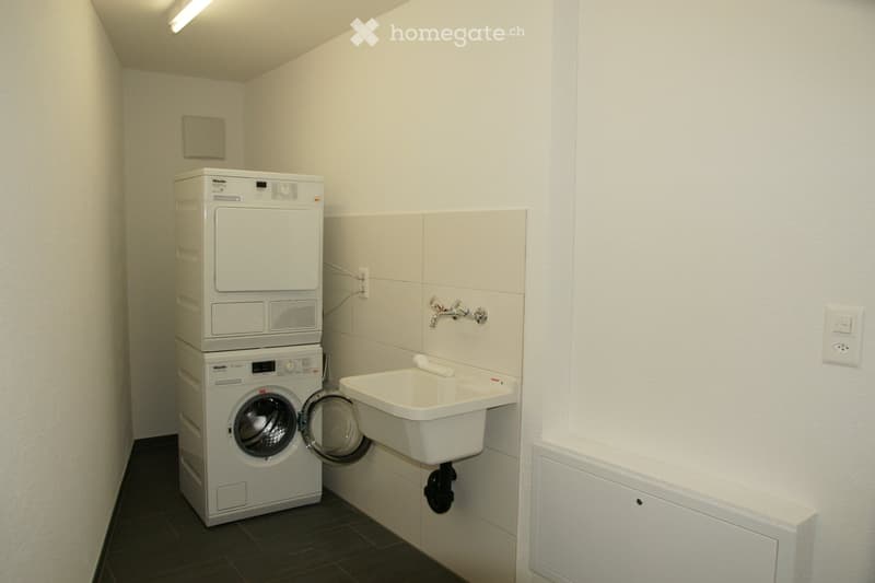 A compact bathroom with a washing machine, a sink, and a faucet, featuring white tiled walls and a dark tiled floor.