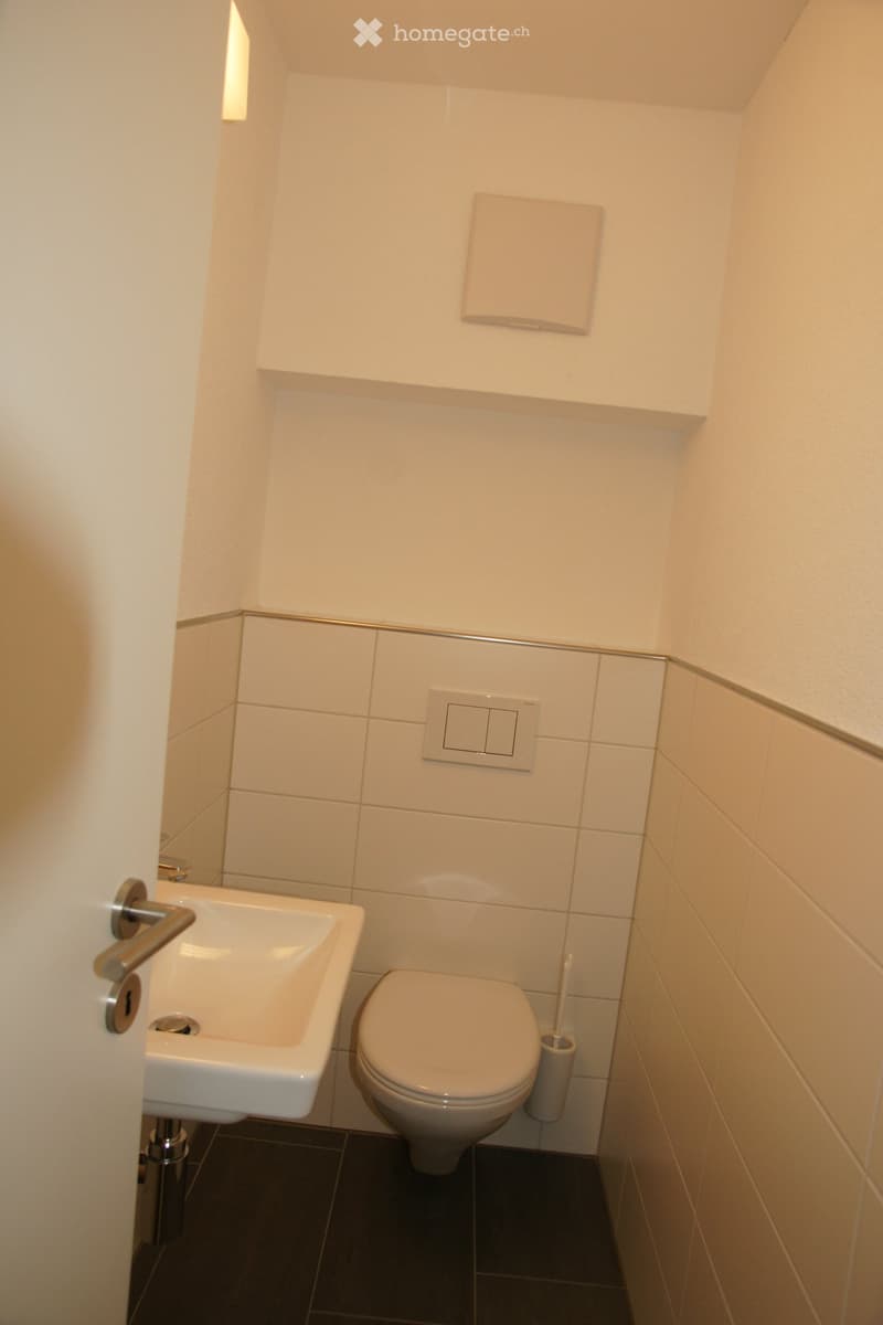 Modern bathroom with white tiled walls, a toilet, a sink, a toothbrush holder, and a towel on a shelf.