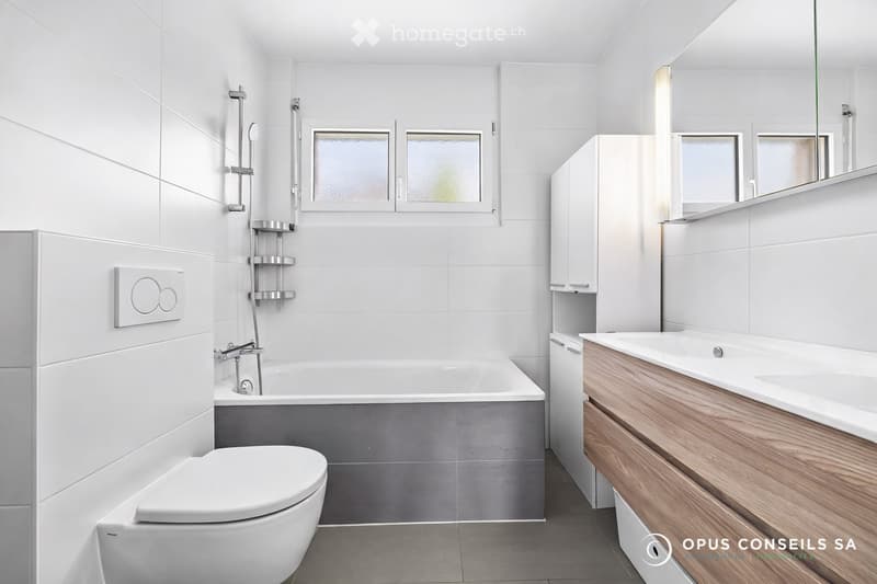 Modern bathroom with white tiles, a bathtub, toilet, sink, wooden cabinet, and a large mirror. Features a shower with a handheld sprayer and a window with frosted glass.