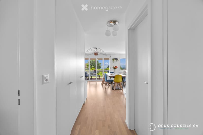 Bright, modern hallway with white walls, wooden flooring, and recessed lighting. Opens into a dining area with a table, chairs, and a view of a garden.