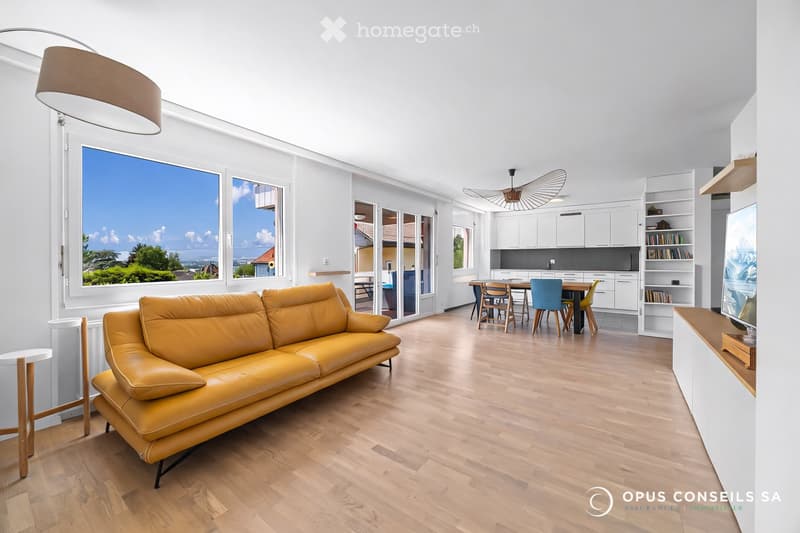 Modern living and dining area with a yellow leather sofa, a dining table with chairs, white kitchen cabinets, and a large window offering a scenic view.
