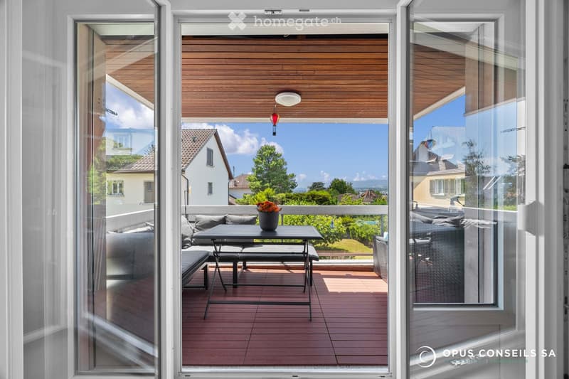 Balcony with wooden decking, glass sliding doors, outdoor furniture including a table, chairs, sofa, and a flower pot, overlooking a scenic view of houses and greenery.