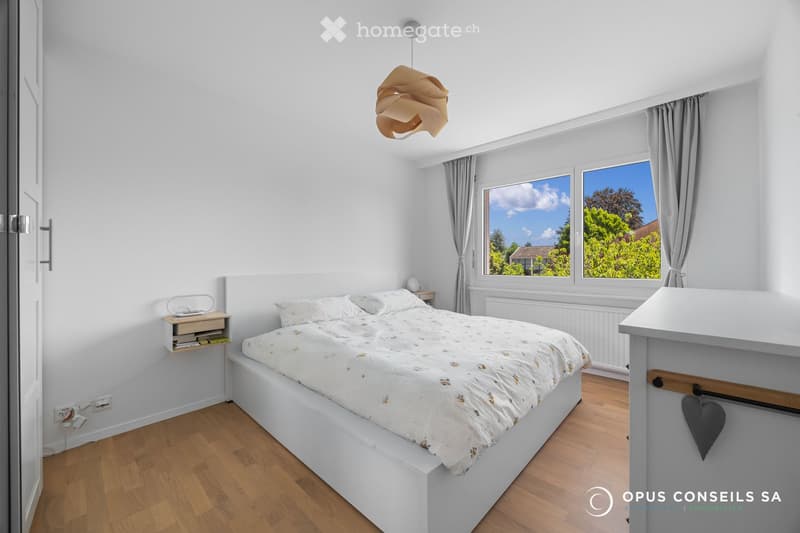 Bright bedroom with white walls, wooden flooring, large windows with grey curtains, a white bed with patterned bedding, a nightstand with a lamp, and a hanging pendant light.