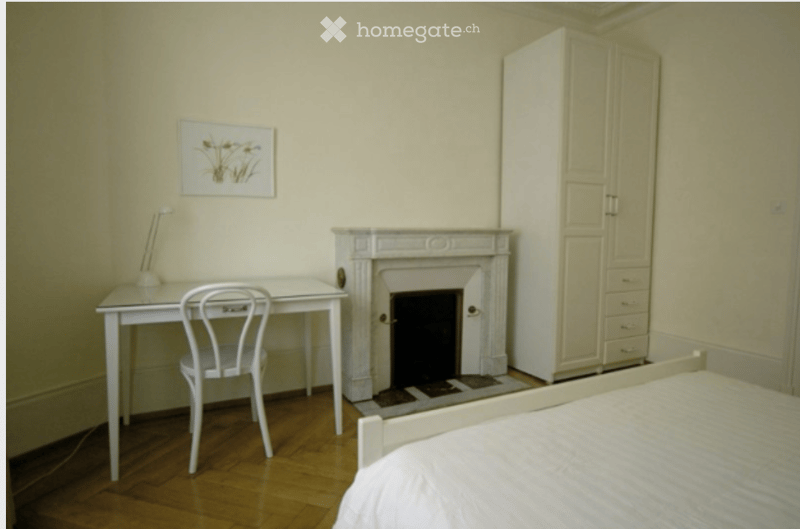 Cozy bedroom with a fireplace, a bed, a white desk with a chair, a wardrobe, and a picture frame on the wall.