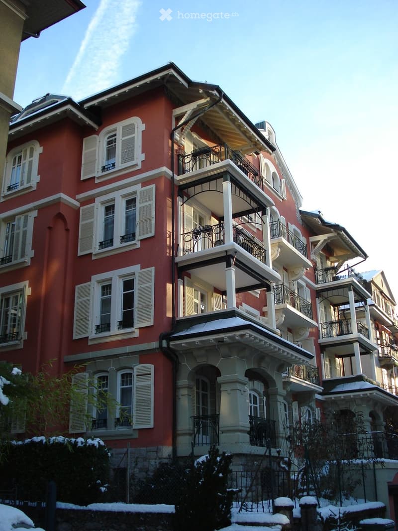 A multi-story red brick building with white-framed windows and balconies, featuring ornate railings and a snow-covered garden area in front.