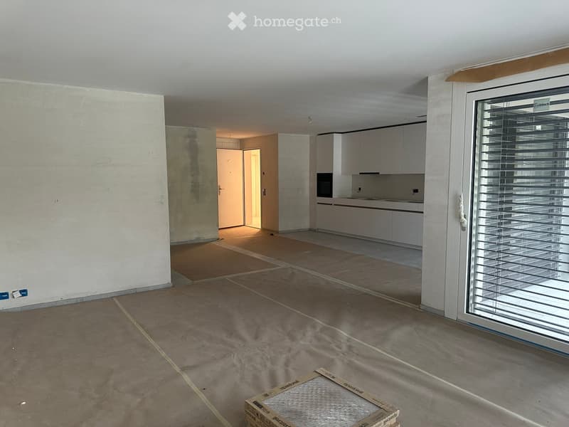 Empty room with unfinished walls and floor, exposed wiring, and a partially visible kitchen area with cabinets and appliances.