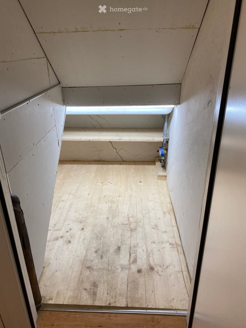 Empty hallway with wooden flooring, exposed beams, and a small window at the far end. The walls are unfinished with visible cracks and patches.