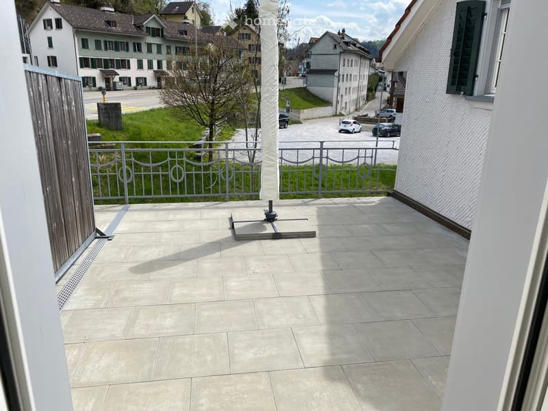 Balcony with tiled floor, metal railing, and umbrella stand, overlooking a residential area with houses and greenery.