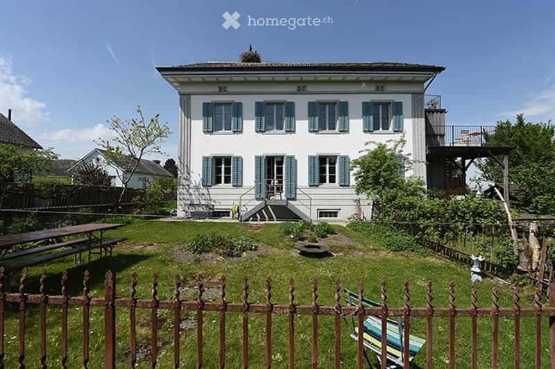 A well-maintained garden area in front of a two-story house with white walls and blue shutters. The garden features a wooden fence, a picnic table, and a small seating area with a bench and chairs.