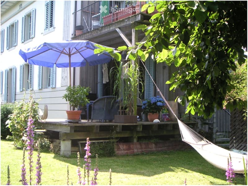 A well-maintained garden with a raised patio area, featuring outdoor furniture, potted plants, a hammock, and a large umbrella providing shade.