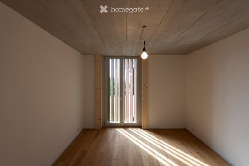 Empty room with light wooden flooring, white walls, and a large window with vertical blinds allowing natural light to enter.