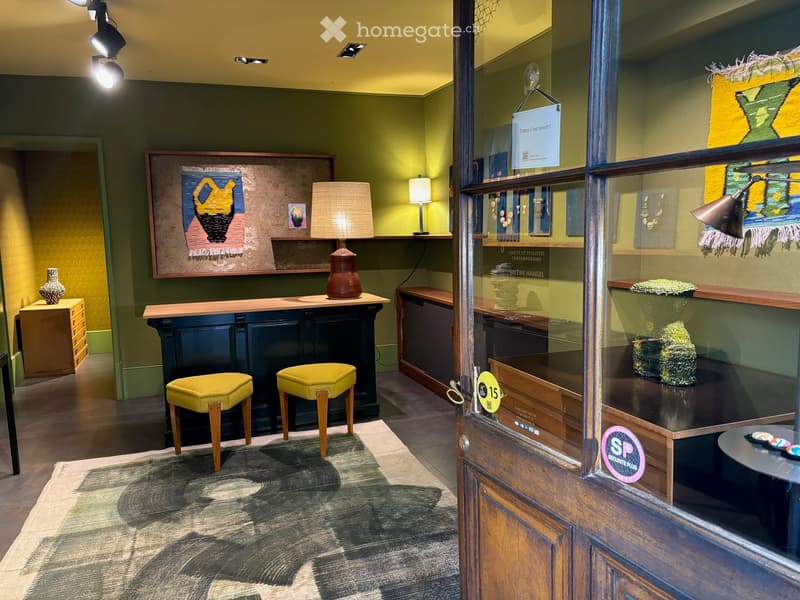 A modern gallery or showroom with a reception area, featuring a reception desk, two yellow stools, a table lamp, a vase, and artwork displayed on walls and shelves.