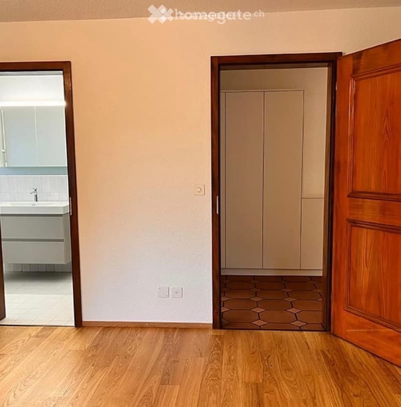 Empty room with wooden flooring, white walls, and a closet with sliding doors. A bathroom is visible through an open doorway.