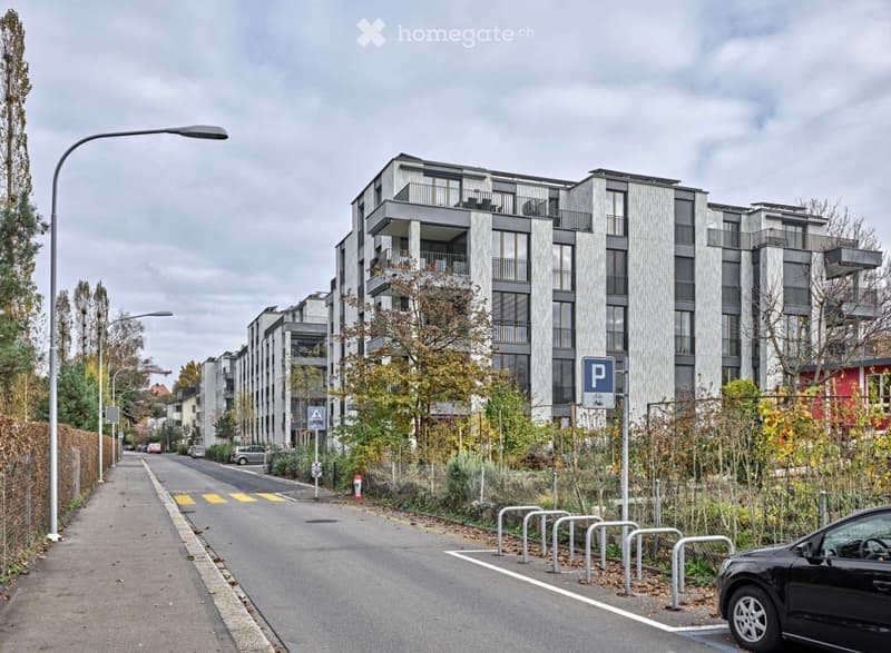 Modern multi-story apartment building with balconies, located on a quiet street with parking spaces and street lamps.
