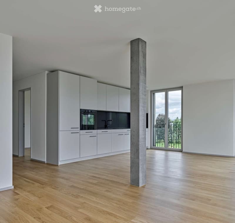 Empty kitchen with white cabinets, wooden flooring, and a large window overlooking a green area.
