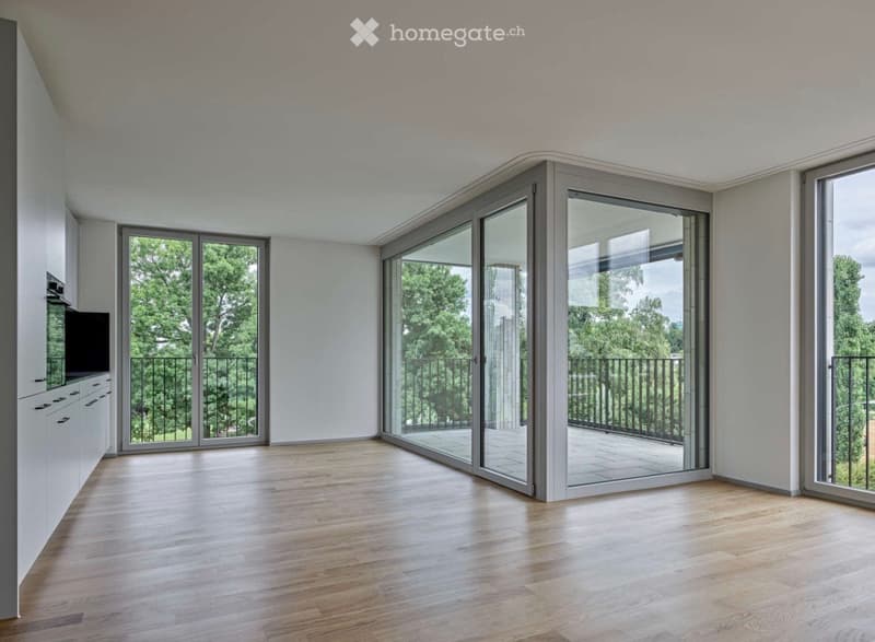 Bright, open-plan room with light wooden flooring, white walls, and large sliding glass doors leading to a balcony with a view of greenery.