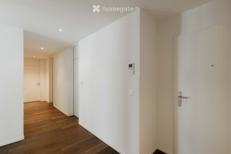 A clean, modern hallway with white walls, wooden flooring, and recessed lighting. Features include a closed door on the right, an intercom system mounted on the wall, and a partially visible adjacent room with a light-colored door.
