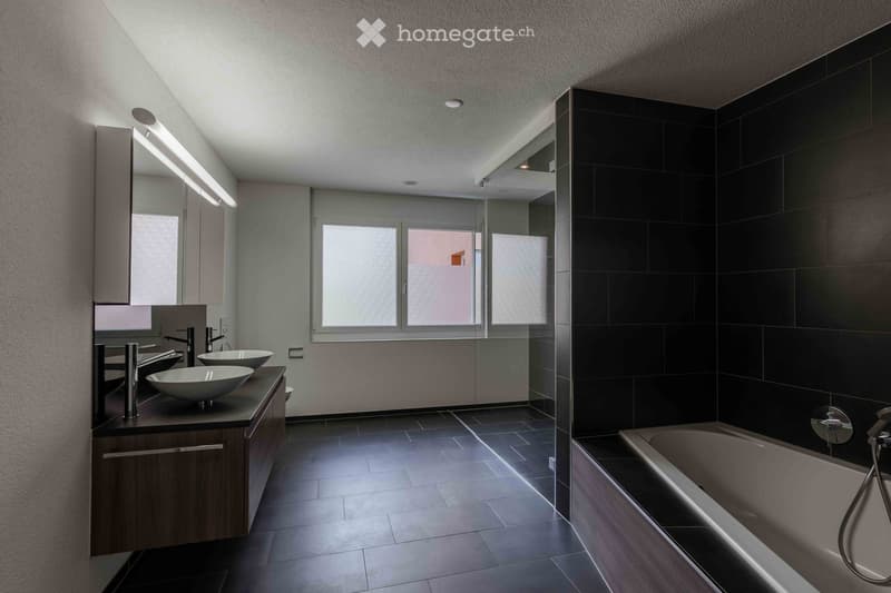 Modern bathroom with dark tiled walls, two sinks, a bathtub, and a large mirror with built-in lighting.